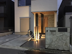 グレー系の天然石を門柱に貼り付け、玄関前には茶系のアルミ角材を建て、ライトを設置/夜間にはライトアップが楽しめる画像